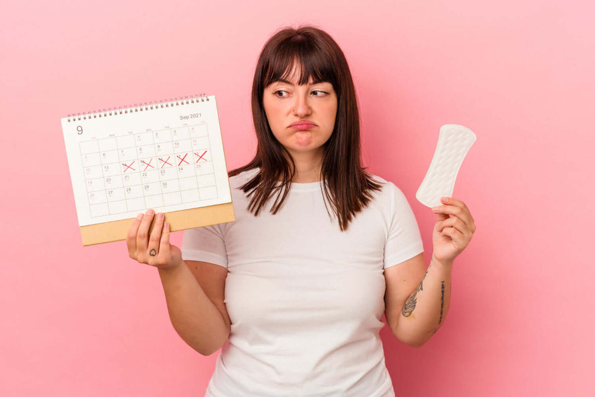 Woman showing an irregular menstrual calendar with a pad, expressing frustration over her period.