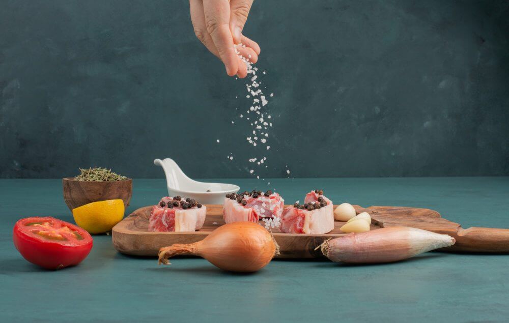 Hand sprinkling Celtic salt over fresh ingredients including tomatoes, onions, and herbs on a wooden cutting board.