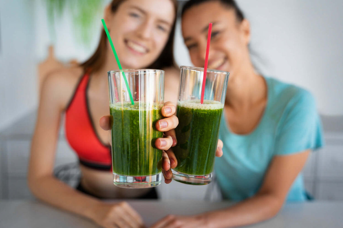 Two women smiling and toasting with green smoothies, promoting health and wellness with greens powder.