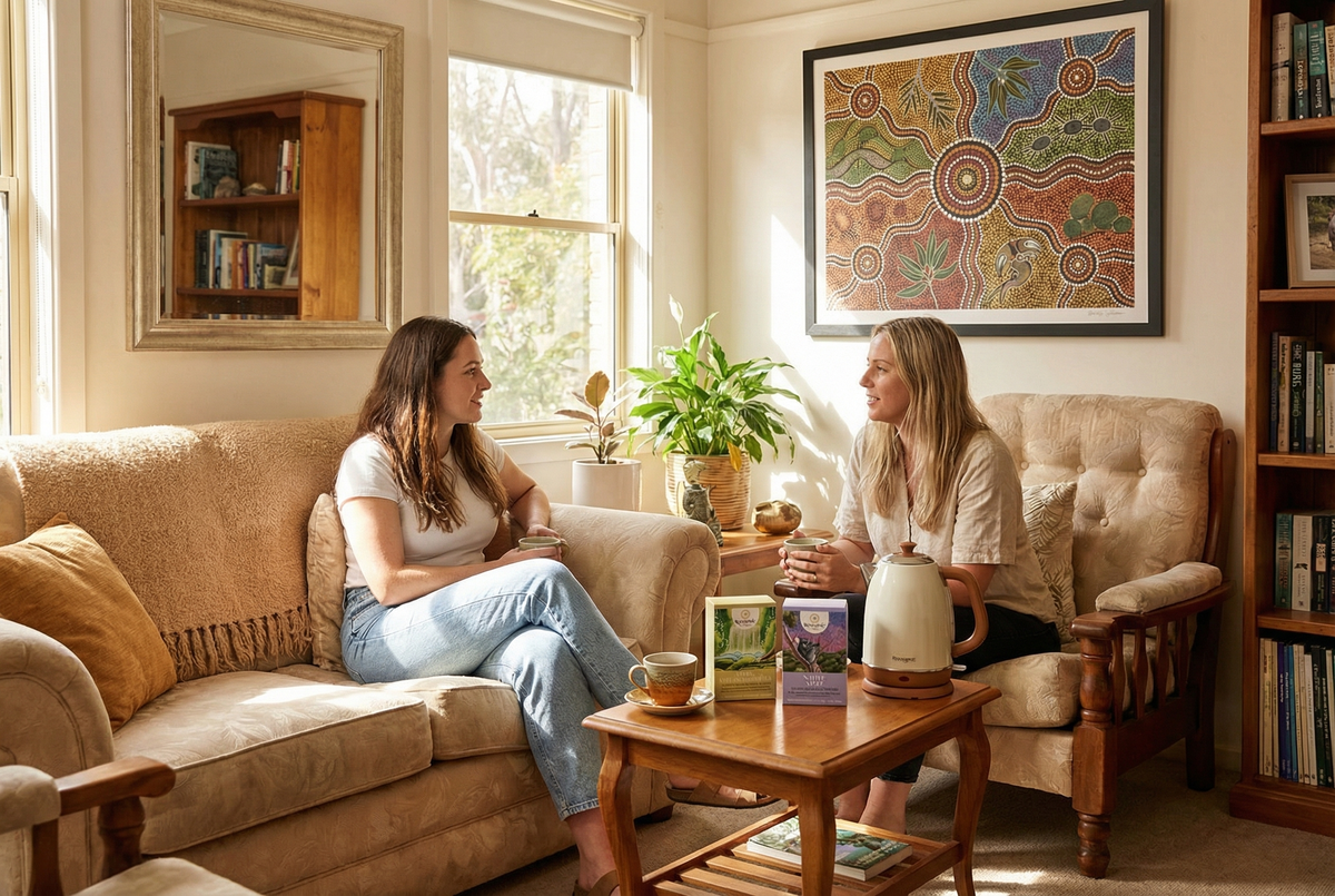 Two women relaxing in a sunlit living room, chatting and holding mugs, seated near a wooden coffee table with a cream kettle and boxes of Roogenic Native Sleep and Detox tea, with a large vibrant Aboriginal dot painting hanging on the wall behind them.