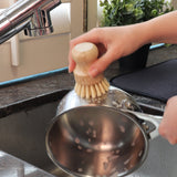 White Magic Eco Basics Plastic Free Pot Brush held in one hand while cleaning a saucepan