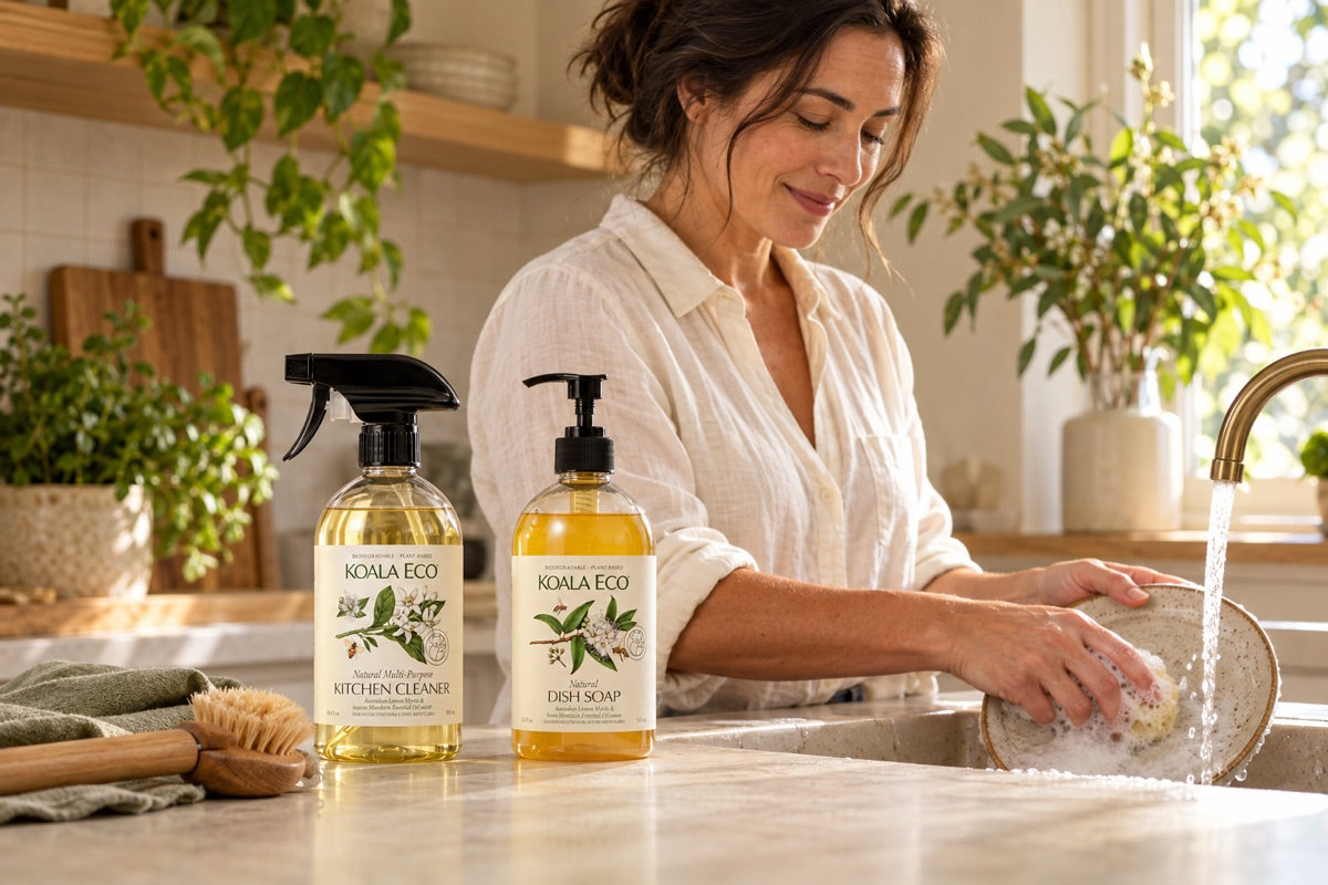 A woman washing dishes in a sunlit kitchen with Koala Eco kitchen cleaner and dish soap on the bench.